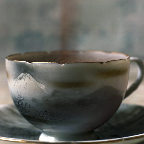 Digital c-print of a photograph which depicts a zoomed in view of an earth toned tea cup with matching saucer. The tea cup is decorated with an Asian watercolor ink landscape of a mountain and several trees. Around the rim of the tea cup there are several evenly spaced indents
