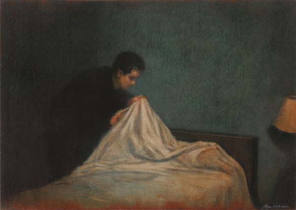 A man examines the sheets of a bed in front of dark blue walls and a lamp.