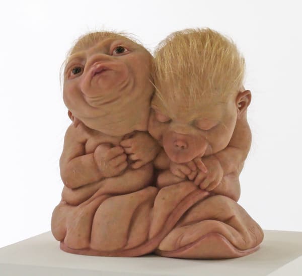 Sculpture of two small hybrid creatures holding each other close. Their faces are like baby monkeys but they have the hands of humans, blonde hair and speckled human-like skin. Their bodies are wrinkled and their backs are like soles of boots.