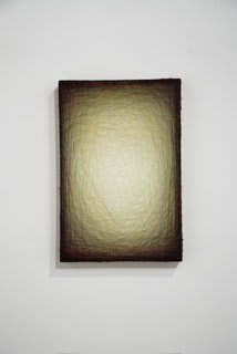 Layers of multi-colored threads strung across a rectangular wooden and plaster panel create the effect of a large glowing light olive green center that is enclosed by a dark brown border