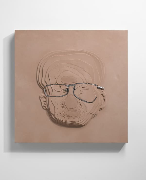 Multiple carvings into beige foam create topographical layers. Warped eyeglass frames are sunken into the center of the foam layers and form an abstract portrait of the artist’s face