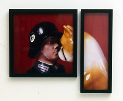A single photograph of a Nazi officer figurine dressed in a balck uniform face to face with a black brown and white dog figurine. The photograph is split and framed into two separate framed pieces with different dimensions