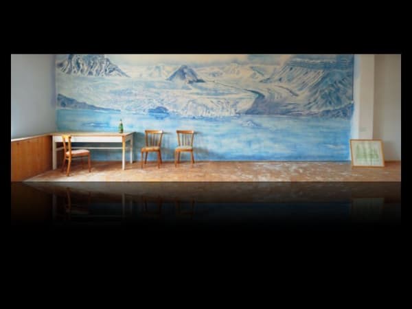Digital panoramic landscape photograph of an abandoned minimally furnished room with a long wall On the long wall a blue toned mural of an mountainous arctic landscape has been painted