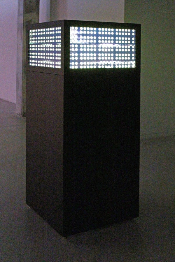 Still from video installation. Tall black box with four screens mounted near the top of each side. The screens show yellow circles organized in columns and rows.