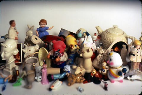 Cibachrome print of a digital photograph which depicts a clustered group of variously sized and different figurines and found objects in front of a white background. A black vignette borders the sides of the print and blurs out some of the objects