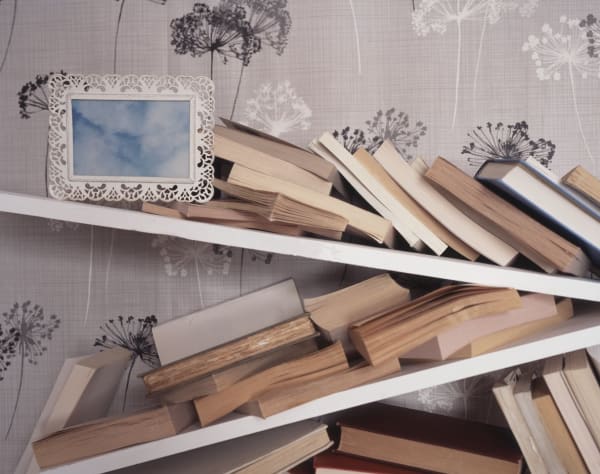 Digital c-print of a photograph which depicts three broken slanted bookshelves against a grayscale floral patterned wallpaper. The bookshelves are covered with toppled and messibly stack yellowed books while at the upper left an intricate lace patterned white photo frame remains perfectly level