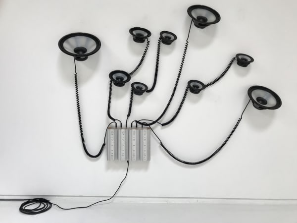 Nine grey and black speakers connected by large black coiled wires mounted on wall in spread out pattern. All cords lead back to silver rectangular box also mounted to wall with single black cable to power source.