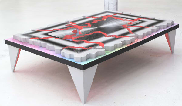 Set atop a pastel rainbow table with sharp points for legs, wood panels are broken into shards. The insides of the panels are painted in red, and the fronts of the panels are painted in shades of black and white rays radiating from the center that are surrounded by concentric rectangles.