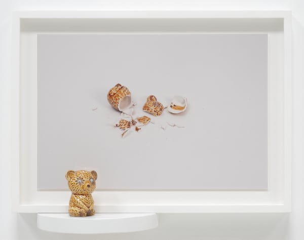 A small teddy bear figurine sits on a ledge next to a framed photo of its shattered body