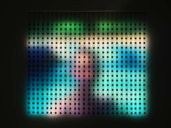 Still image of multiple large hanging multi-colored LED strips attached to a long horizontal metal pole at the top The LEDs light the wall behind them with blurred scenes taken from old family home movies