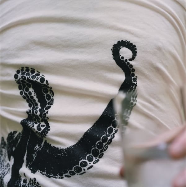 Digital c-print of a photograph which depicts a zoomed in view of the torso of a person wearing a white t shirt with a black octopus printed on it. Blurred out in the foreground is the person holding a glass of water