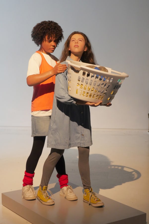 Sculpture of two lifelike young girls in casual clothing holding laundry basket with towels and sleeping furry creature wearing gloves. Girls are mid-walk and one girl grips the shoulder of the girl in front of her.