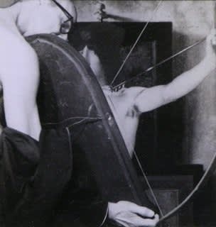 Black and white scene with artist’s self portrait viewed from the back holding a long oval shaped frame. Behind the artist’s self portrait is a shirtless man with his arms outstretched.
