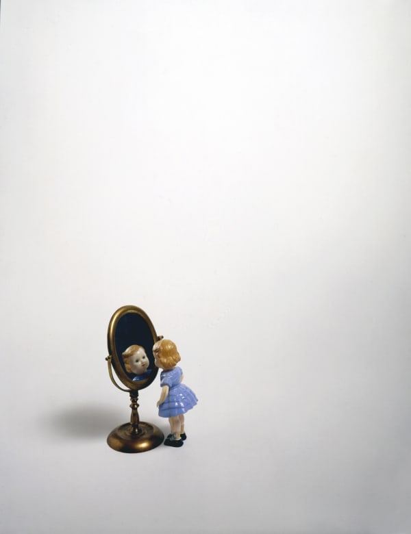 White sheet with doll figurine looking into tiny mirror reflecting face.