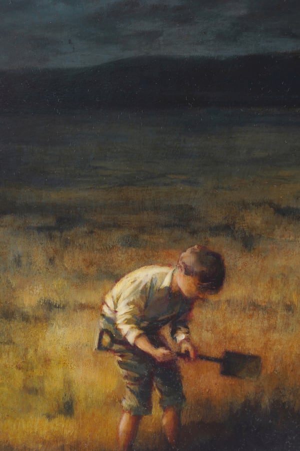 A child holding a shovel leans over in a field of yellow grass with mountains and a dark sky in the background.