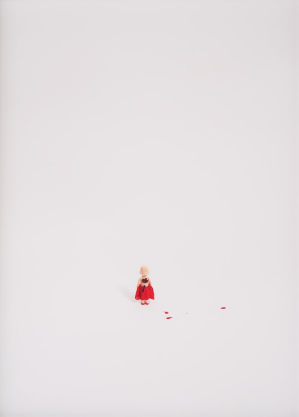 A photograph of a figurine of a girl dressed in red with an axe and splatters of red paint against a white background.