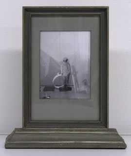 Black and white scene of person wearing shapeless jumpsuit and standing in room filled with assortment of shapes and objects.