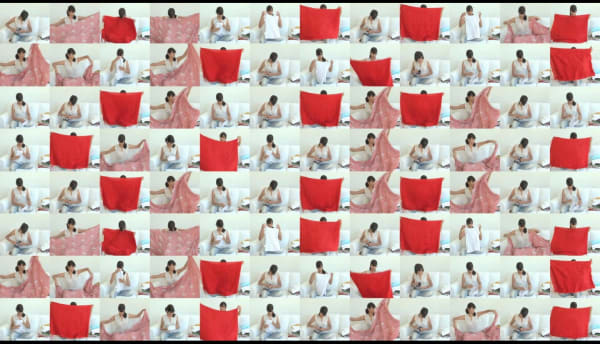 96 video frames arranged in an eight-by-twelve grid. In each frame, Surabhi Saraf is seated on a white couch folding three large sheets (one white, one patterned and pink, one red). The video plays on the frames in a coordinated manner to create patterns of movement within the grid.