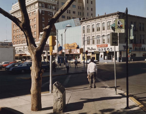 man in white shirt standing at crosswalk with tree in foreground and stores on other side of the street, by Stephen Shore