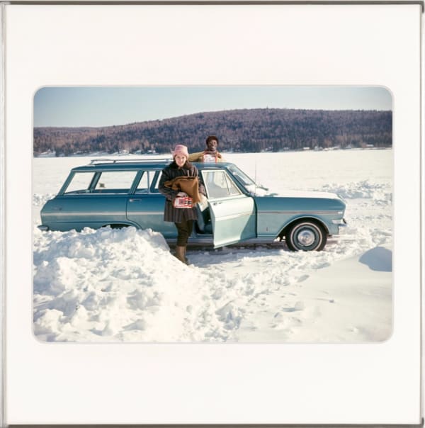 Omar Diop standing by the driver's door of a car looking over the hood while a white woman stands in the passenger door holding a case of beer. Both are looking at the camera. The car is parked in a snow field. By Lee Shulman & The Anonymous Project.