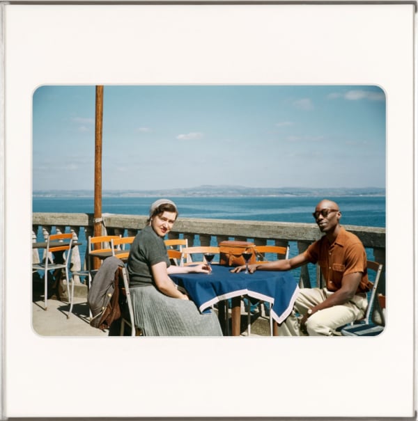 Kodachrome slide of white woman and artist, Omar Diop at table with scenic sea view. By Lee Shulman & The Anonymous Project.