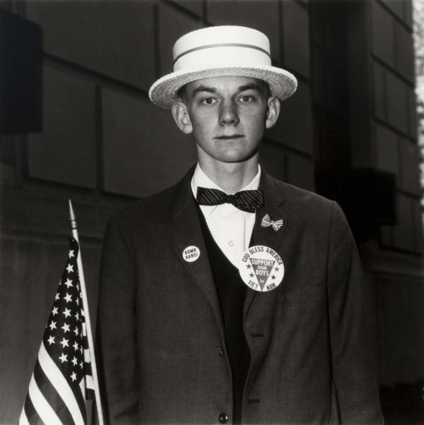 Diane Arbus portrait of young white man in straw hat, waiting to march in a pro-war parade in NYC, holding an American flag, with Bomb Hanoi and Support Our Troops pins
