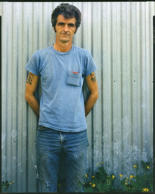 Bruce Wrighton Man with tattoed arms, in blue t-shirt, in front of green corrugated fence, yellow flowers at bottom Tirage C-print d'époque 20 x 25 cm Dim. papier: 20 x 25 cm