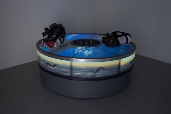 Sandra Ramos Perpetual movement of the Cuban Worm between two shores (uniformes), 2018-2023 Lightbox. Kinetic Installation. (A baggage carousel and primary school uniforms typical of Cuban schools, packed in transparent briefcases) Height: 29 in Diameter: 81 in