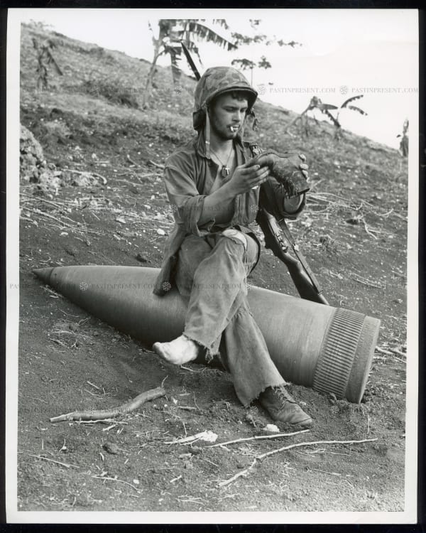 A. B. Knight, “At Ease” Using an unexploded 16 inch naval shell for a resting place, Marine Private First Class Raymond L. Hubert, shaken a three-day accumulation of sand from his boondocker Saipan Marianas 7.4.44, 1944