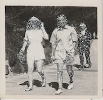 John Latour, Couple dressed in shorts walking alongside a road while a man in black follows behind – reading a book. A second woman trails in the distance, 2020