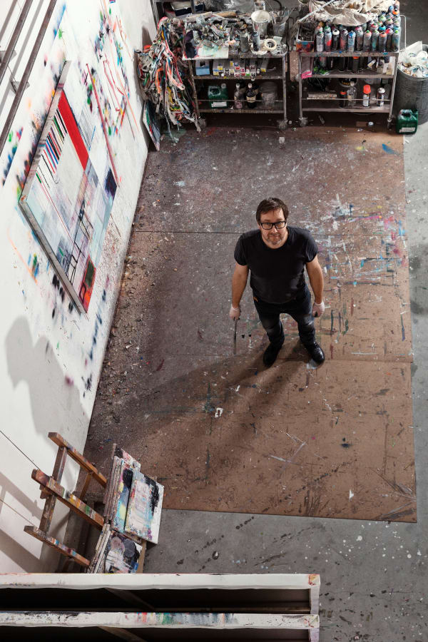 Morten Lassen looks up at the camera in the studio while creating an abstract and geometric painting that uses complex colours and textures.