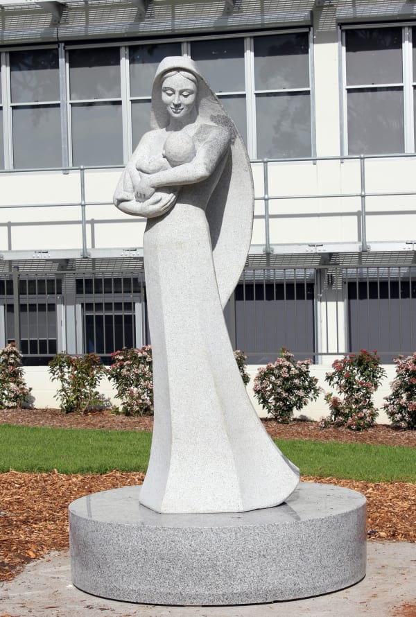 A minimalist granite statue of a woman holding a baby, with a long flowing robe and circular base, set in front of a modern building and manicured lawn.