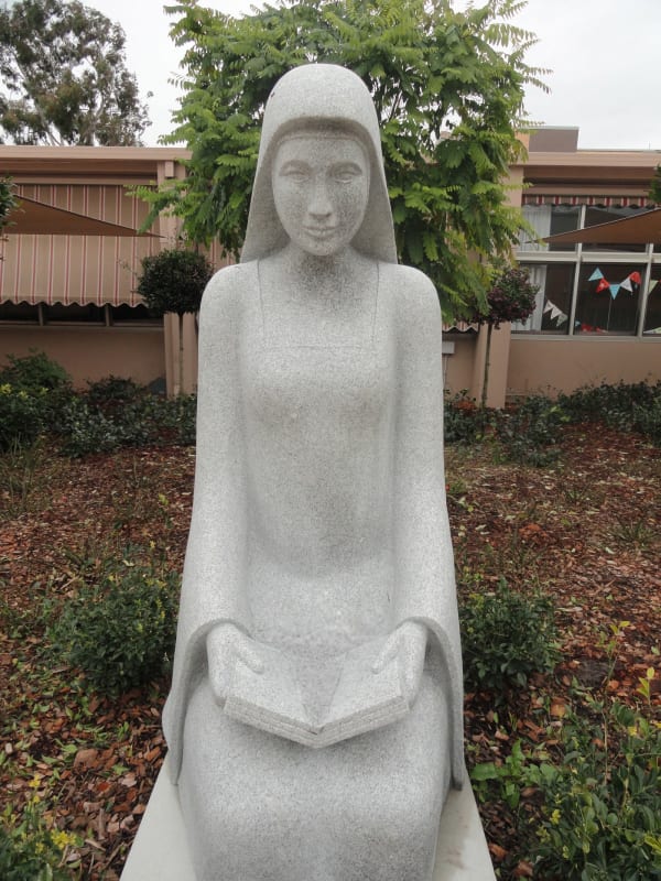 A front-facing granite statue of a serene woman in a simple, flowing dress holding an open book on her lap, surrounded by shrubs and a building in the background.