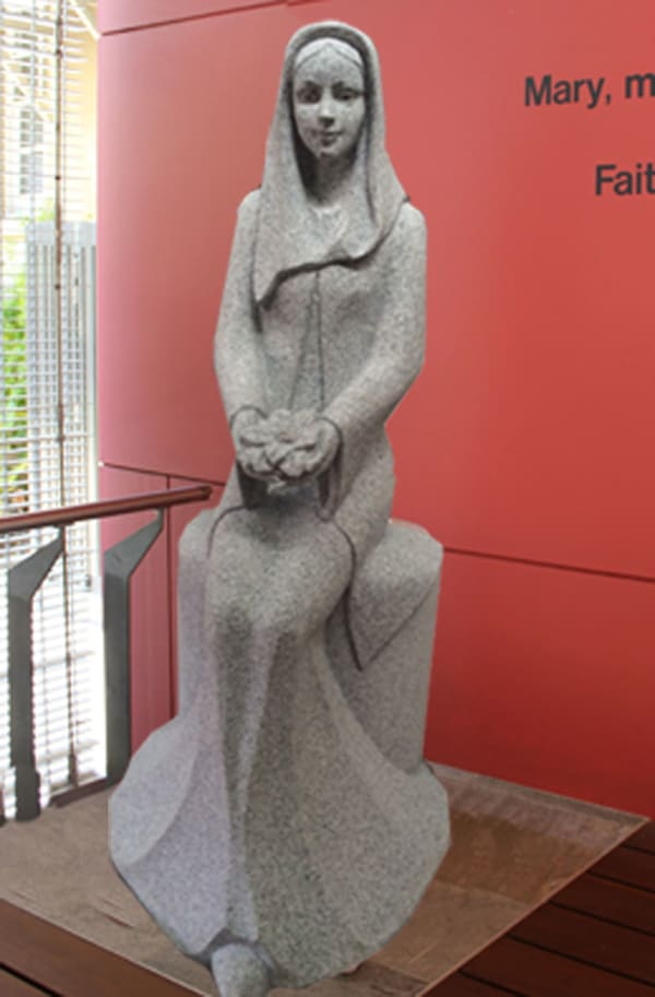 A granite sculpture of a seated woman with cupped hands holding a magnolia, positioned against a bold red wall with the words "Mary, mother of Jesus" partially visible.