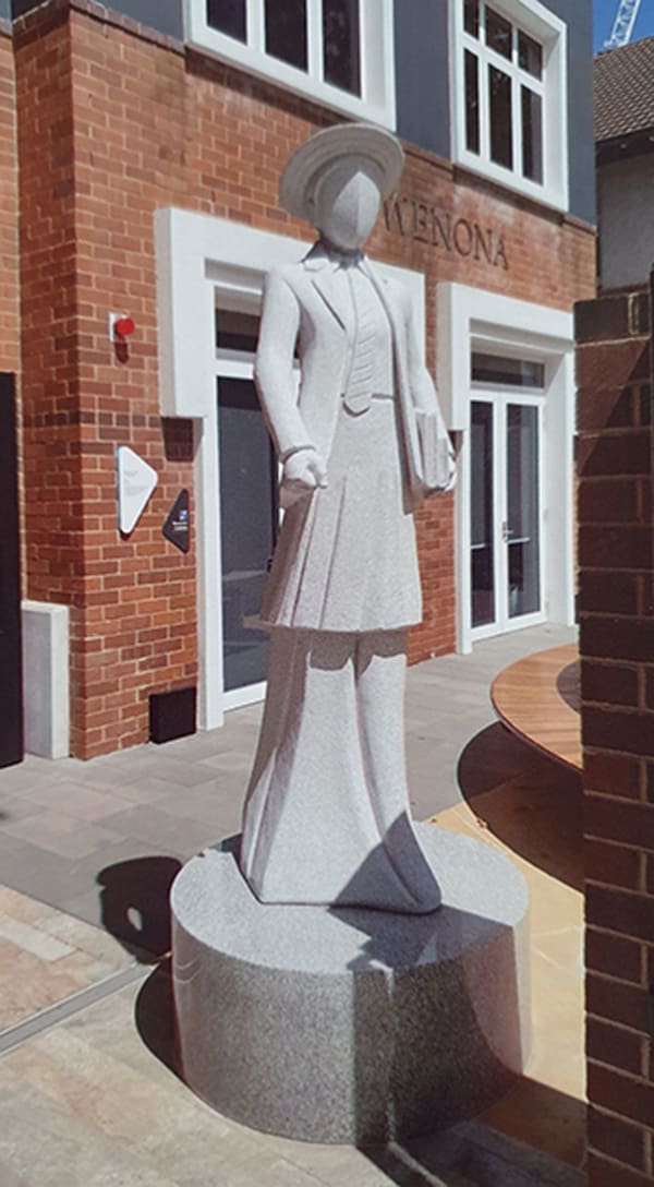 A modern granite statue of a faceless female figure in a school uniform, wearing a brimmed hat and holding a book. The statue is positioned in front of a brick building with "WENONA" engraved on the wall.