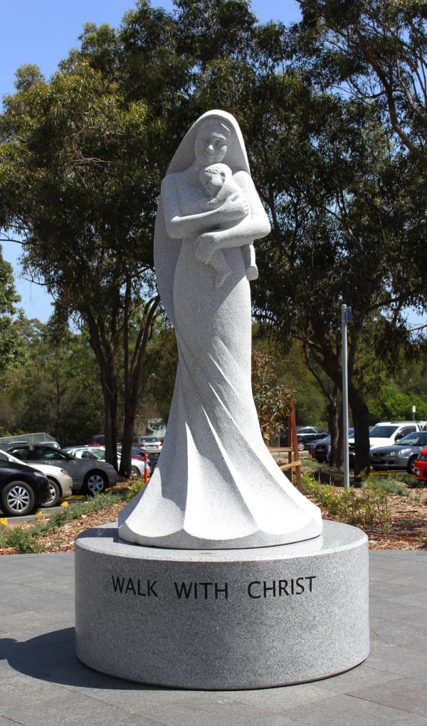 A smooth granite statue of a serene woman holding an infant close to her chest, standing on a circular base with the words "Walk With Christ" engraved. The statue is surrounded by parked cars and lush trees.