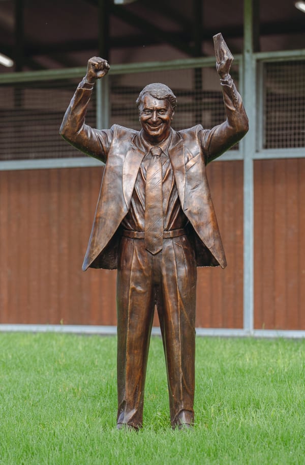 A bronze statue of Nick Columb standing on a green lawn, with both arms raised in a victorious gesture. He is depicted in a suit and tie, with an exuberant expression, holding a book in one hand. The statue is set outdoors, with a wooden stable-like structure in the background, symbolizing his achievements in the horse racing industry.