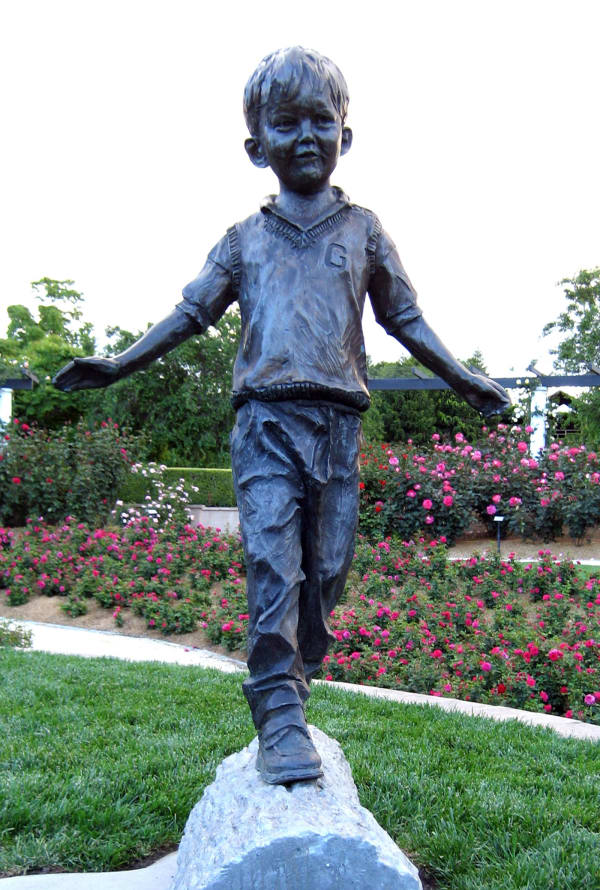 Bronze sculpture of a young boy balancing on a log, part of a whimsical children's garden scene in a lush park setting.