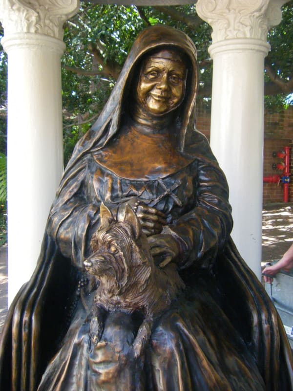 Close-up of a bronze statue of St. Mary MacKillop seated with a gentle smile, holding her beloved dog, Bobbs, in her lap. The finely detailed sculpture captures her warmth and compassion.