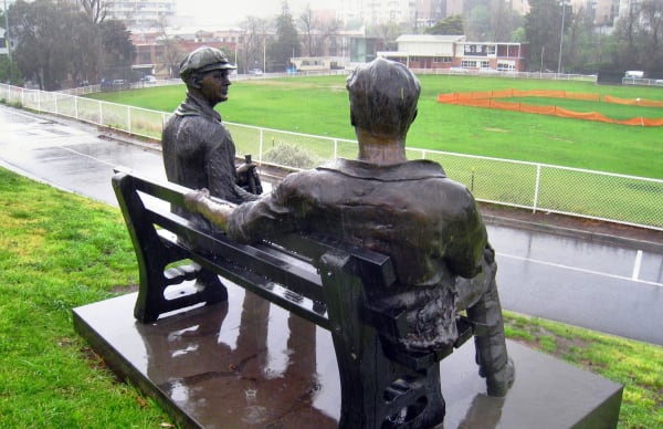 Bronze sculptures of cricketers Bill Woodfull and Keith Miller seated on a bench, overlooking a cricket field. The figures, depicted in casual poses, evoke a sense of camaraderie and nostalgia in a rainy outdoor setting.