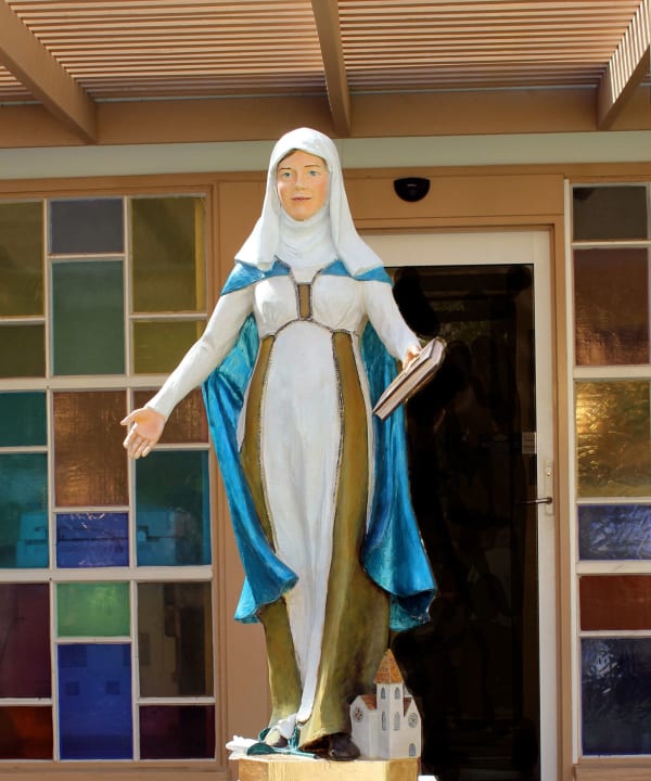 Resin statue of St. Catherine of Siena, depicted in a white habit with a blue mantle, holding an open book in one hand and extending the other in a welcoming gesture. The sculpture stands in front of a building with colorful stained-glass windows, symbolizing her role as a scholar and spiritual guide.