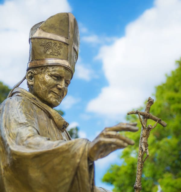 Detail of a bronze sculpture of St John Paul II holding a staff topped with a crucifix.. The scene is set outdoors on a sunny day, with a tree in the background.