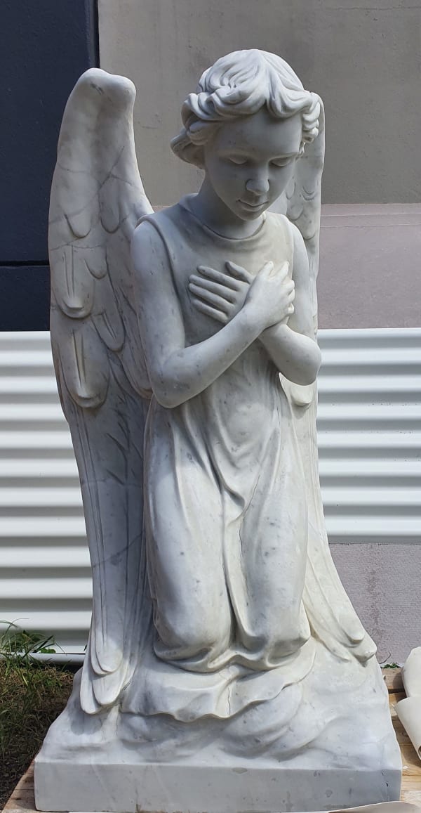 A full view of a kneeling marble angel statue with intricately carved wings, hands crossed over its chest, and a flowing robe. The statue captures a moment of quiet devotion, standing against a plain backdrop.