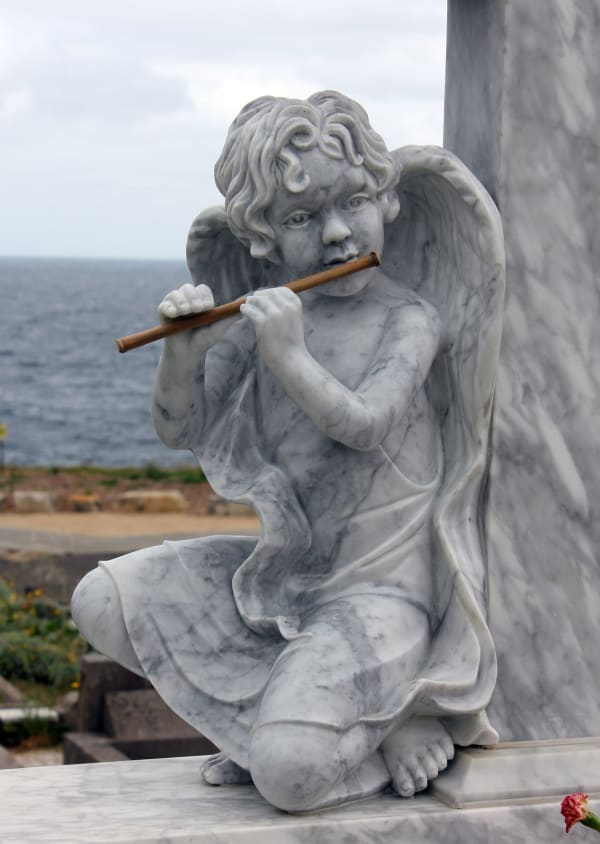 A marble statue of a cherubic angel kneeling and playing a flute. The soft folds of the robe and textured curls of the hair add life-like realism, with an oceanic backdrop enhancing the serenity of the scene.
