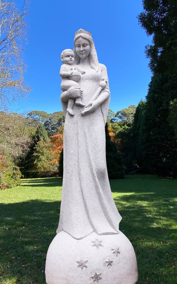A tall granite statue of a crowned woman holding a baby, standing on a spherical base adorned with engraved stars. The statue is placed outdoors in a green park with a clear blue sky.