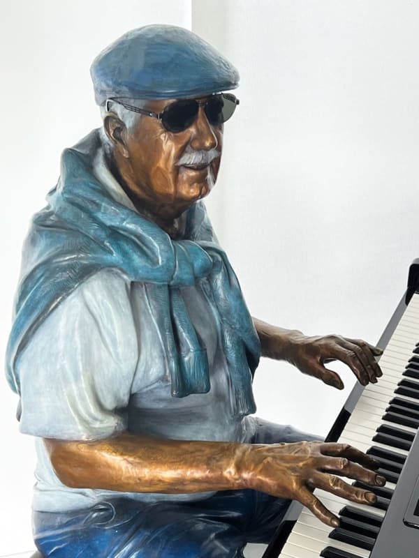 Detail view of upper torso and hands of the bronze sculpture of an elderly man seated at a grand piano, playing with a serene and focused expression. The figure is dressed casually in shorts, a t-shirt, and a sweater draped over his shoulders, with vibrant color accents highlighting his clothing and shoes. The artwork captures a moment of musical engagement and nostalgia in a modern indoor setting.