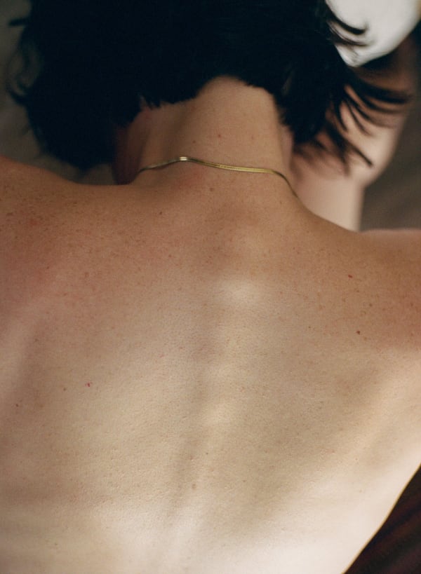 Photograph by Casey Joiner of a woman's lightly sunned and freckled back. Her spine is accentuated. She has short dark hair and wears a gold chain.