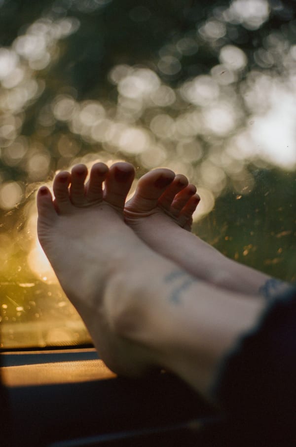Photograph by Casey Joiner of a close up of two feet pressed against glass ( seemingly a windshield). There is a tattoo visible on each foot, and the two feet together block a source of light that shines on them. The blurry background beyond the glass is green and yellow in hues and is reminiscent of nature.