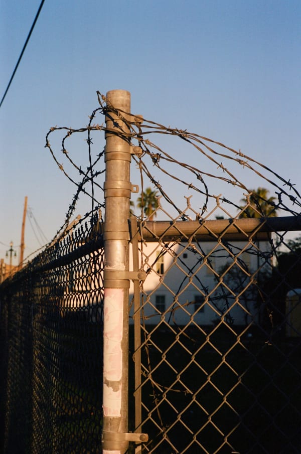 Photograph by Casey Joiner of the corner of a chain linked fence with loosely wrapped barb wire around the pole and along the top. The background is mostly in the shadows thought the sun illuminates the fence, and the sky is very present with it's brilliant blue ombre.