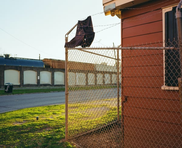 Photograph by Casey Joiner of the corner of a red house with a chainlink fence and haphazardly installed barbed wire with a piece of fabric caught and tangled in it. The background reveals the street (Tchoupitoulas St) and a train peeking over a wall. The grass between the house and the street is illuminated by the evening sun and the sky is warm but pale blue.